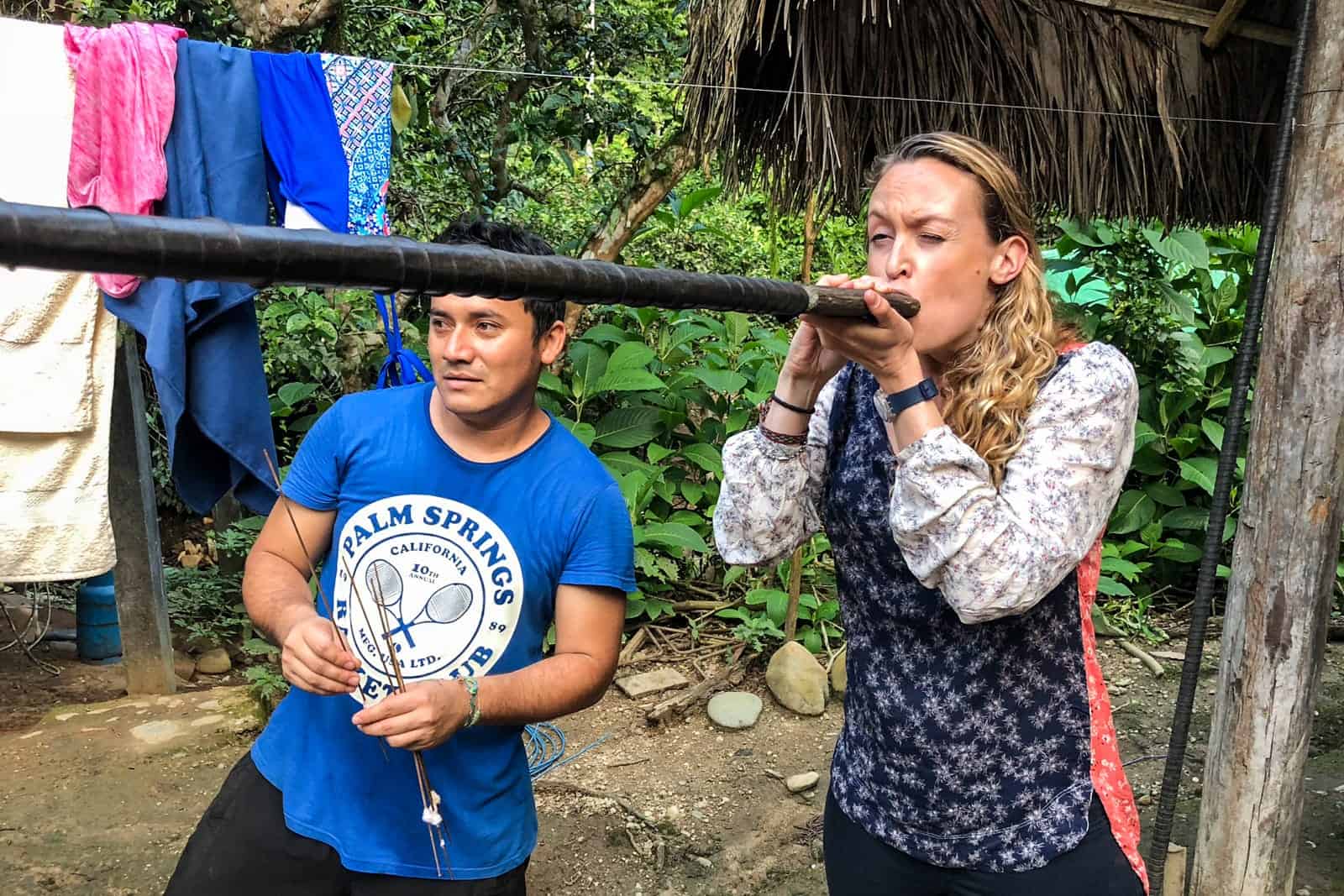 A woman blowing into the black tube of a Blowgun in Ecuador Amazon Rainforest, while a man in a blue t-shirt next to her watches the target. Behind them is a clothes washing line. 