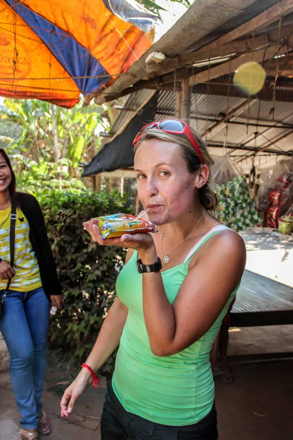 A woman in a green vest drinks a bagged drink from one hand at a stall in rural Cambodia. 