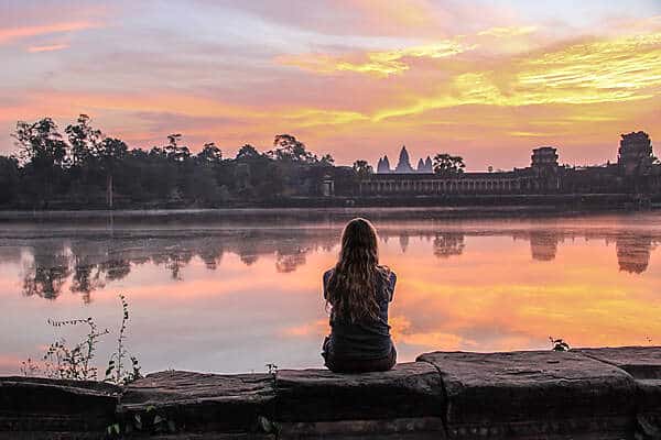 A woman sitting on a stone wall looking towards the complex of the Angkor Wat temple at sunrise. 