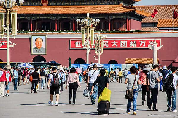 People walking towards the Forbidden City palace whose red walls feature Chinese symbols and a portrait of Chairman Mao. 