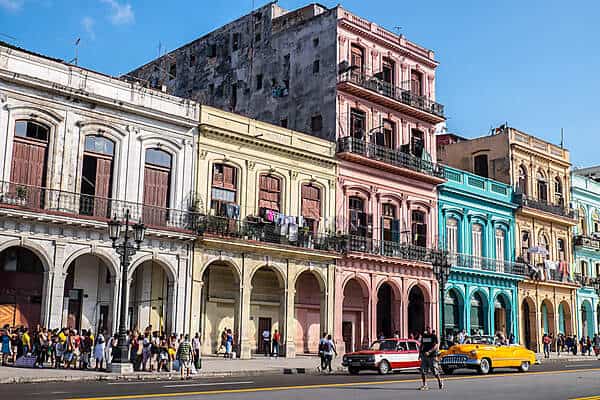A cluster of people and two old cars outside a row of classical pastel coloured buildings in Havana, Cuba. 