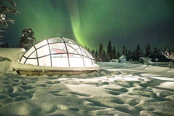 A glass igloo on deep snow under the night sky of green northern lights. 