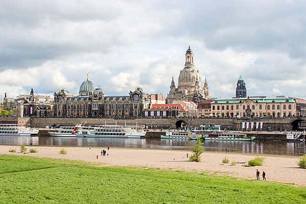 View from a park to a stretch of long, grand, domed buildings along a boat-lined river in Dresden.