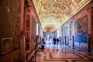 People walking past the delicate mural walls and under the golden arched ceiling Inside the Vatican Museum hallways in Rome