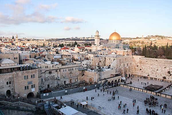 People gather in the Western Wall Plaza in Jerusalem, surrounded by light orange stone walls beneath the golden dome of Temple Mount and a minaret. 