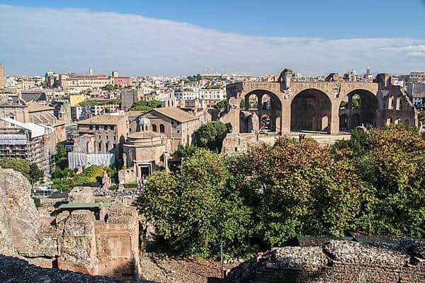 The archway columns, old houses, towers and crumbling walls of the tree-filled ancient ruins of the Palatine Hill in Rome. 