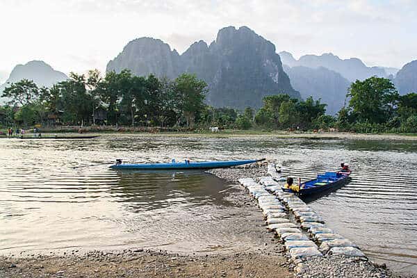 Two blue longboats on a still river in Lao backed by jagged rock formations. 