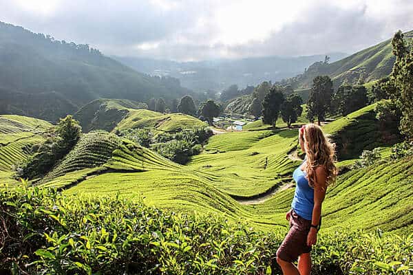 A woman with long hair stands in front of a landscape of rolling green terrain and forested hills. 