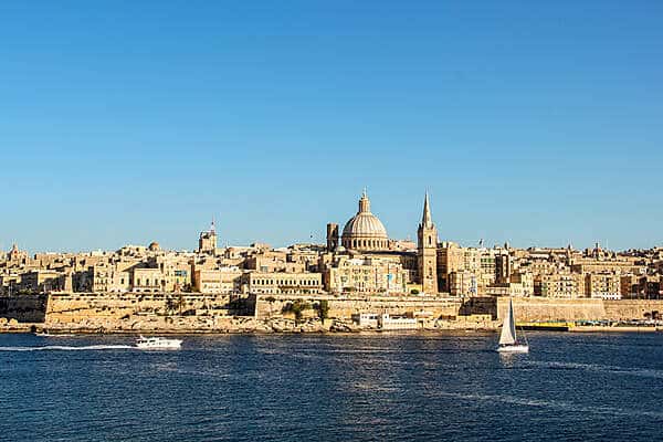 Far view to the golden structures, spires and domes of Valletta, Malta next to the sea, where boats are sailing. 