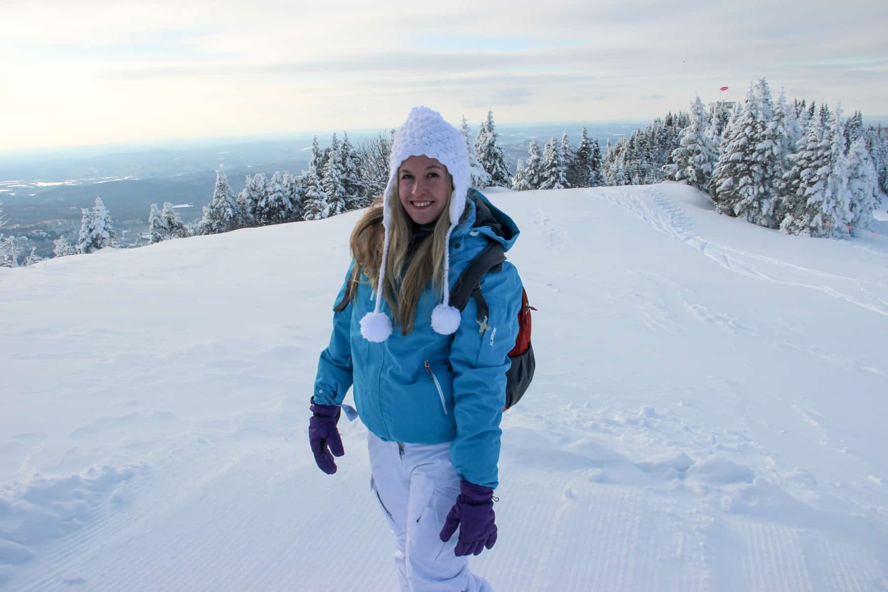 A woman in a blue ski jacket and white trousers and bobble hat stands on a snow covered mountain slope in Mont Tremblant in winter. 
