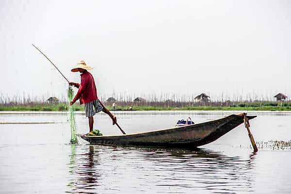 A man in a red shirt and white hat balances on a boat in Inle Lake with one leg, using the other leg to move the paddle.