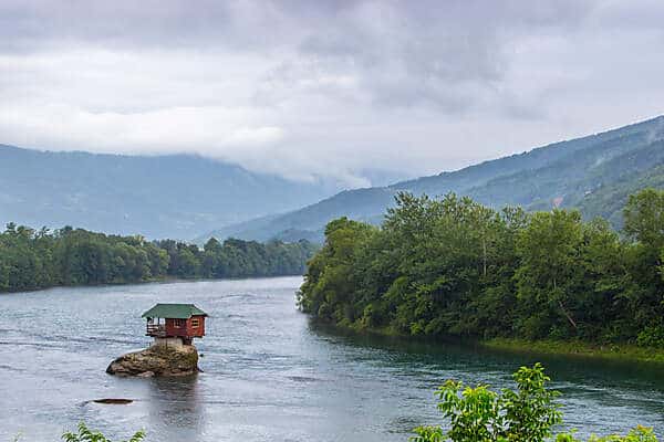 A tiny red house in Serbia, perched on a rock in the middle of a river, surrounded by forest. 