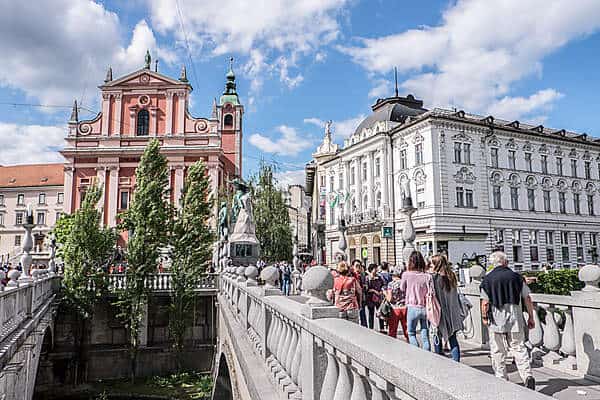 People walking over a triple bridge towards pink and white classical buildings in Ljubljana, Slovenia. 