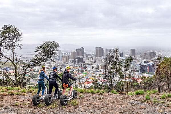 Three women on scooters on a mountain ledge overlooking the city spread of Cape Town, South Africa.