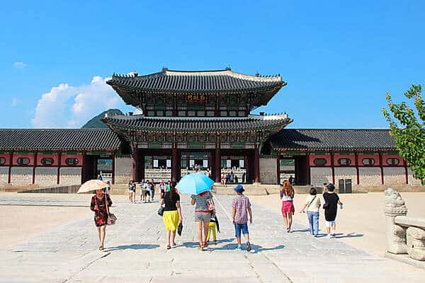 People with sun umbrellas walking towards the two-tier pagoda of Gyeongbokgung Palace in Seoul, South Korea. 