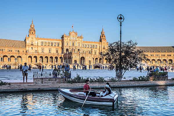 In Seville, a man rows a boat with two passengers on a small manmade river in front of a colossal golden building that glows in the sun.