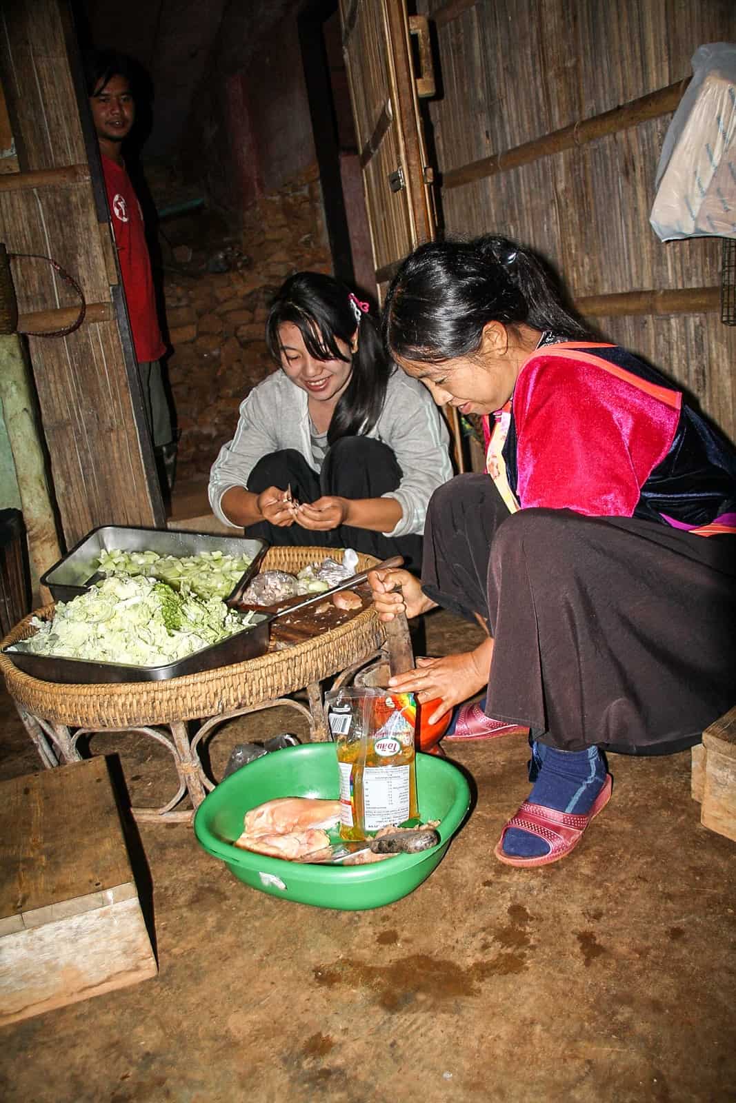 A man looks towards two Thai women crouched and cooking a meal with vegetables and meat in a wooden house. 