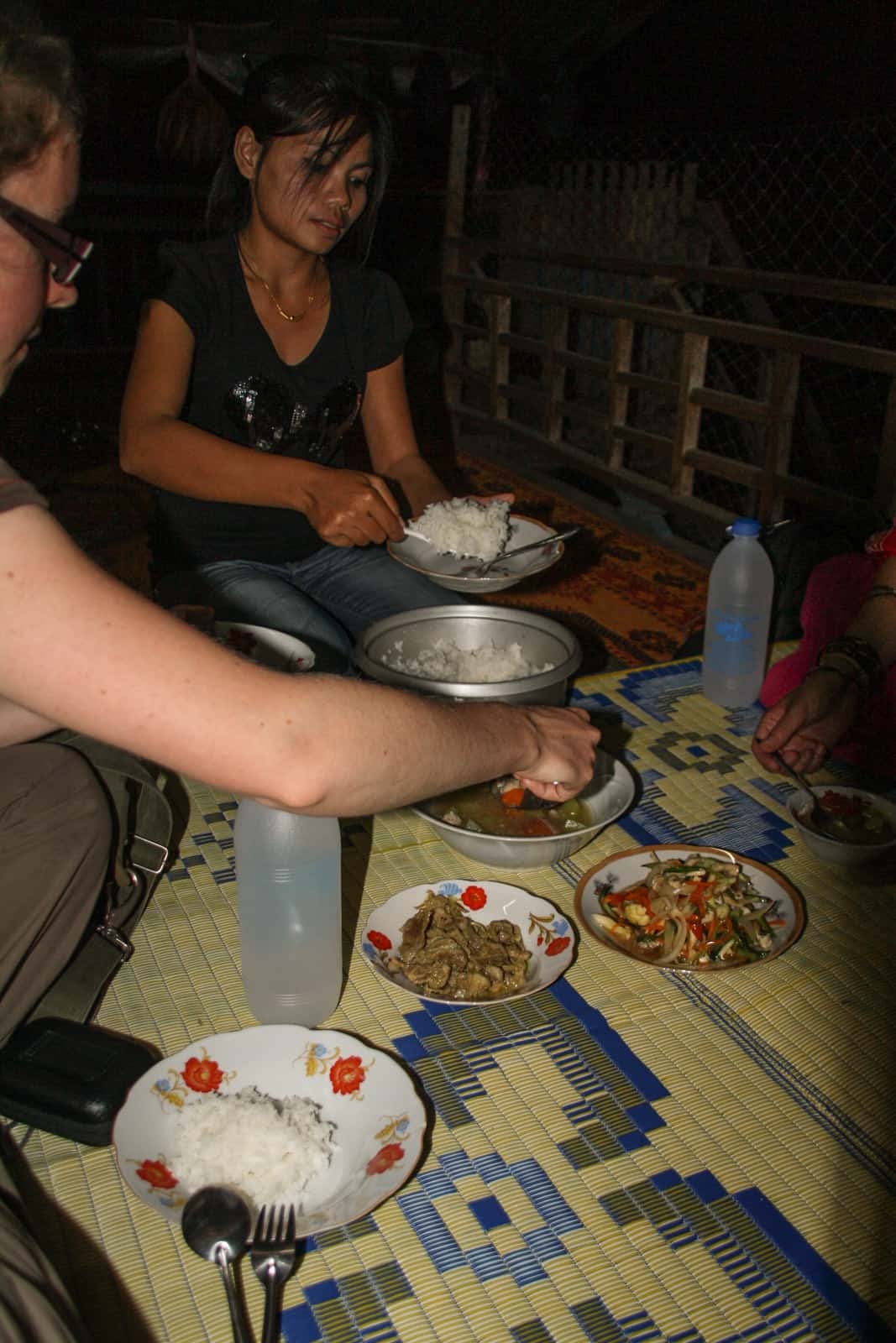 A woman sits on a bamboo mat sharing food dishes with a local Laotian woman. 
