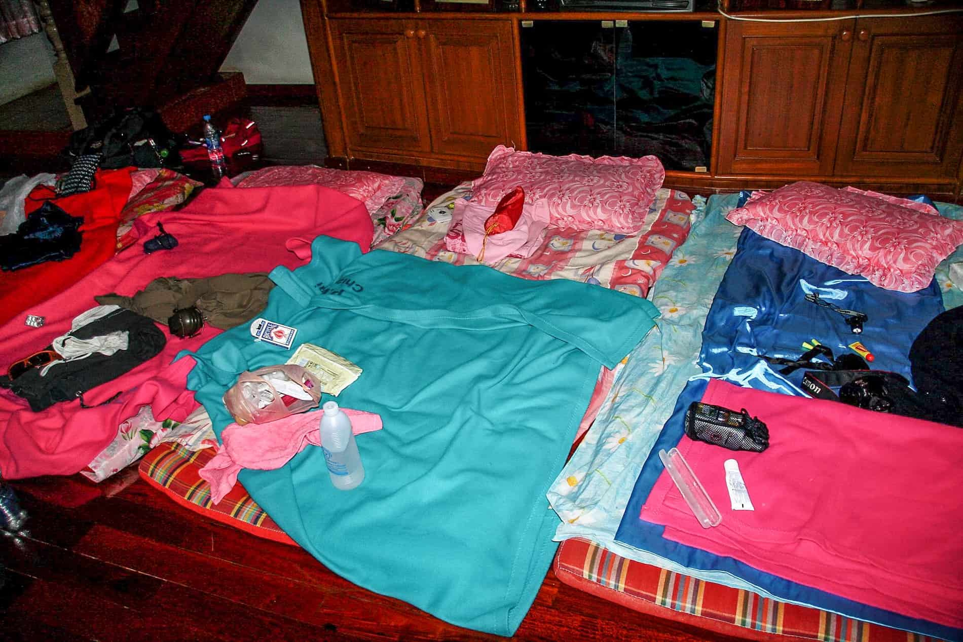 A row of sleeping mats with pink and blue blankets, strewn with travel gear and amenities, on a floor in a house in Thailand. 