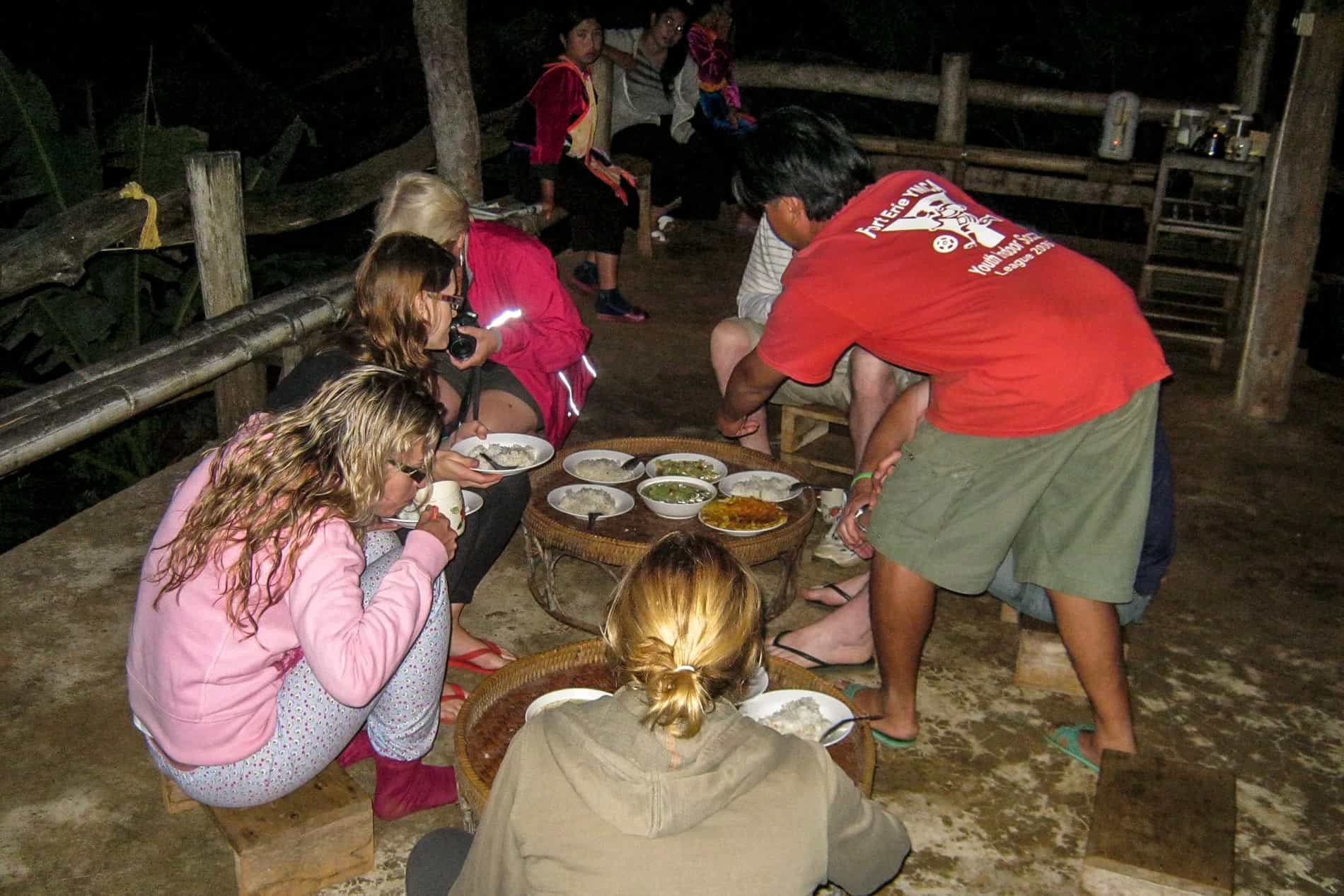 People sitting on low stools and gathered around a table with bowels of food in a homestay in Thailand. 