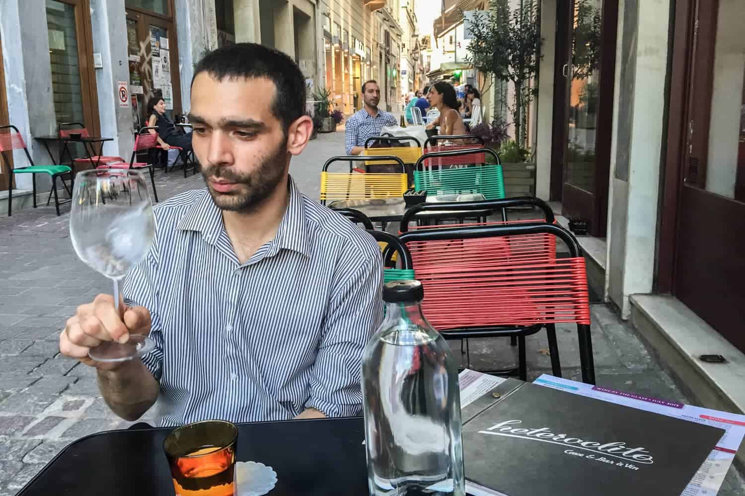 A man looks at a glass of wine while seated at a black table at a wine bar in Athens with red, green and yellow chairs behind him. The black menu next to him bears the bar's name: Heteroclito. 