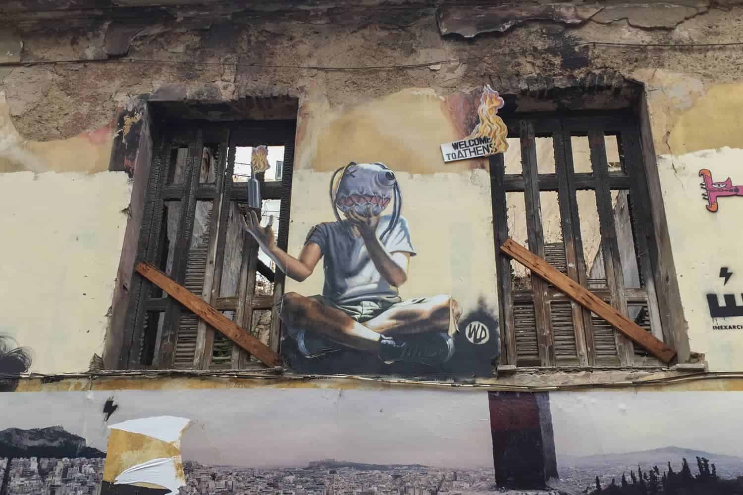 Athens street art on an abandoned building showing a guy in a mask, sitting crossed legged holding a molotov cocktail. Below is a panoramic photograph of the city. 