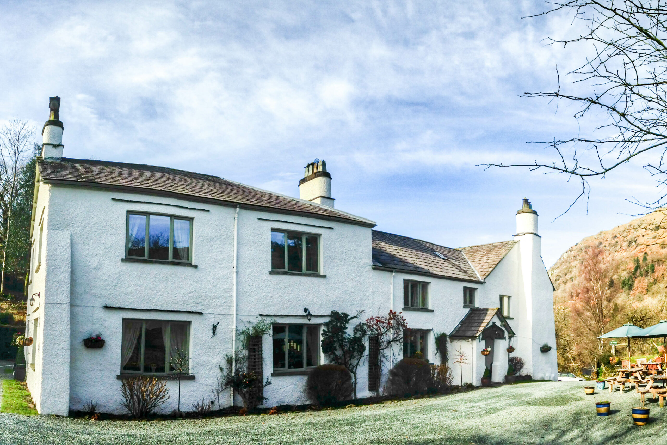 A white cottage with green window trims set within a garden and surrounded by orange foliage hills.