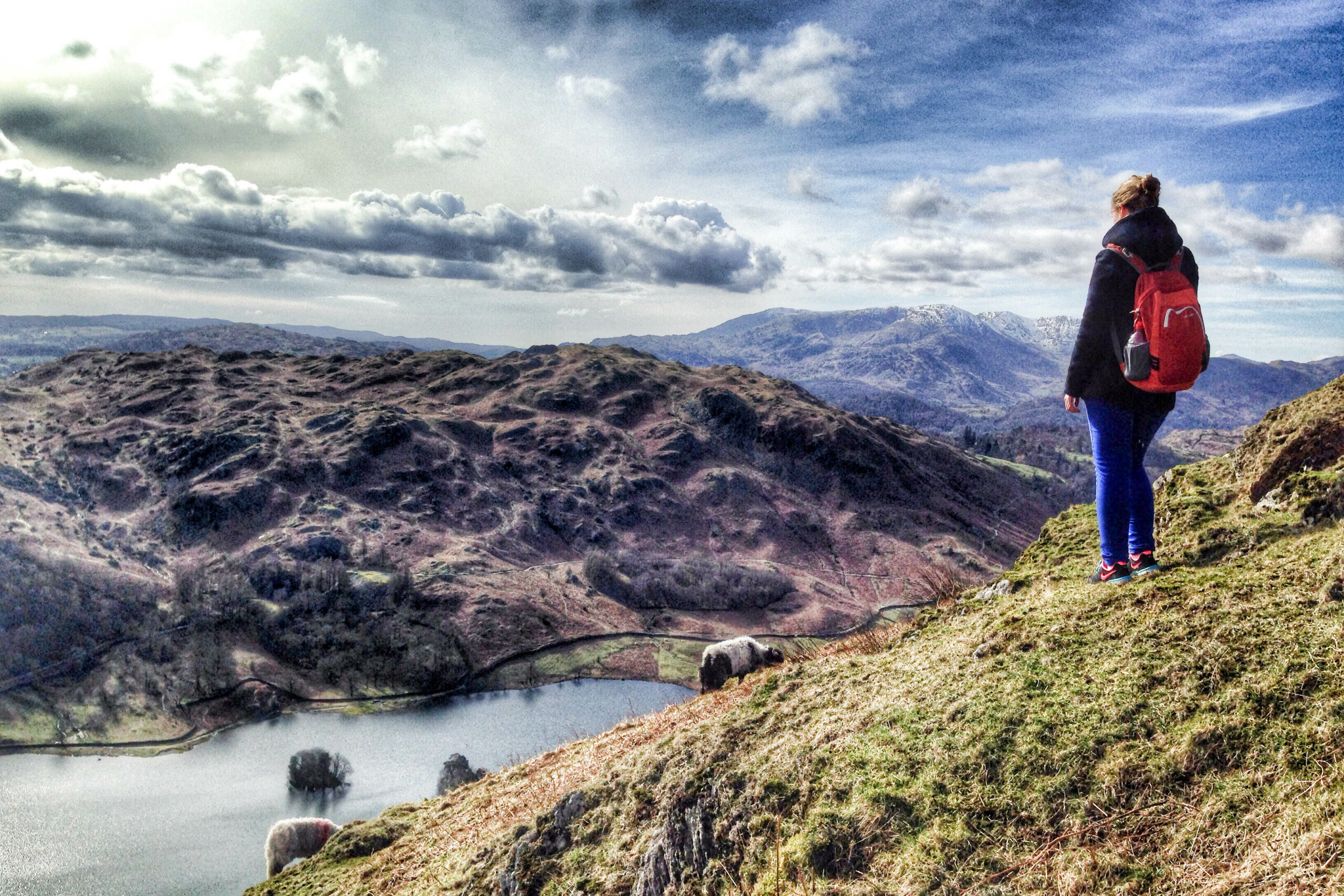 A woman in dark blue clothing and wearing a red backpack, stands on an elevated green grass hill overlooking a lake backed by rugged hills.