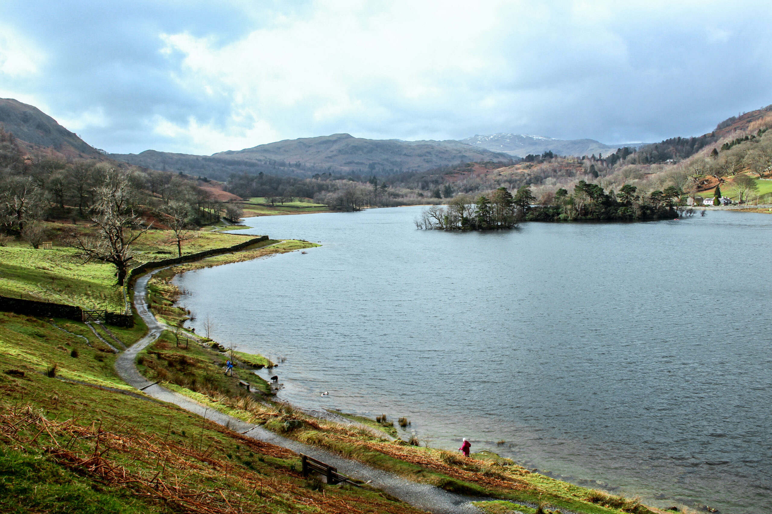 A path on the Fairfield Horseshoe Hike through green land, next to a wide expanse of lake with a small tree island in the middle.
