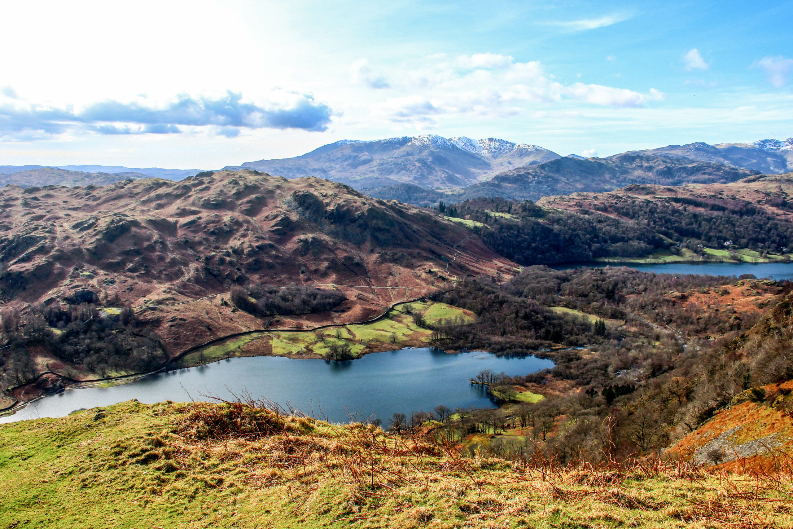 An elevated view from a grassy mountain ledge over a dusky blue lake backed by ochre soil and foliage covered hills.