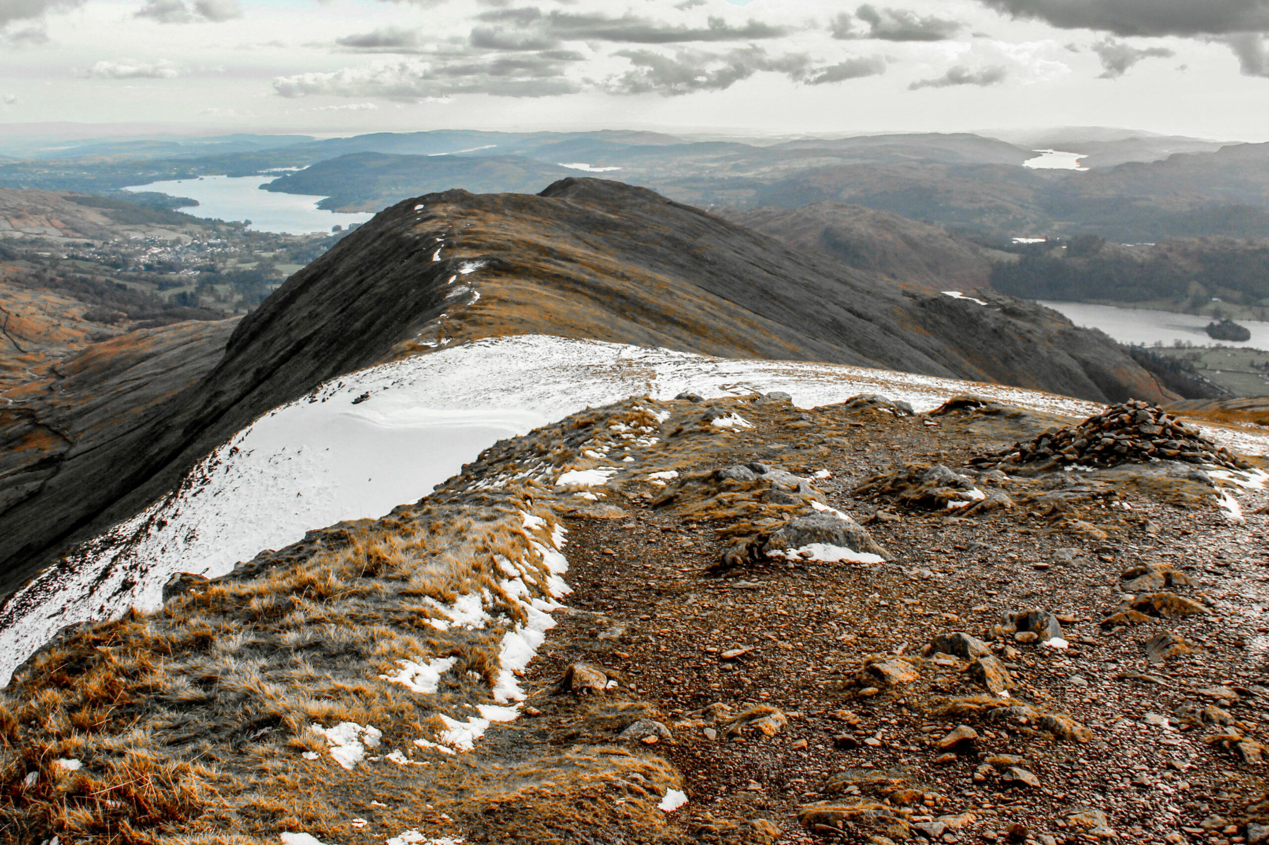 View from a rocky, snow-covered hilltop towards an undulating landscape and a far-distance lake.