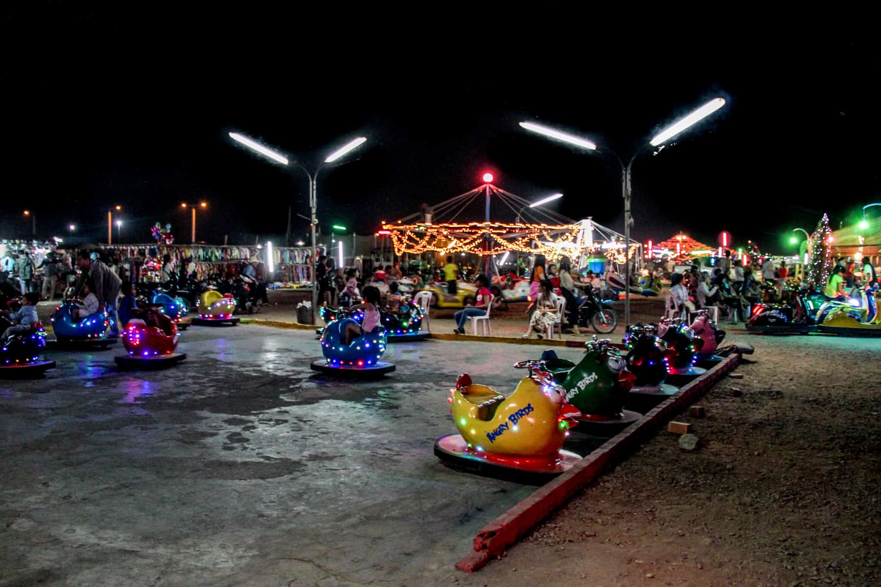 A fair ground scene at night with children's bumper cars in the foreground and merry-go-round rides in the background. 