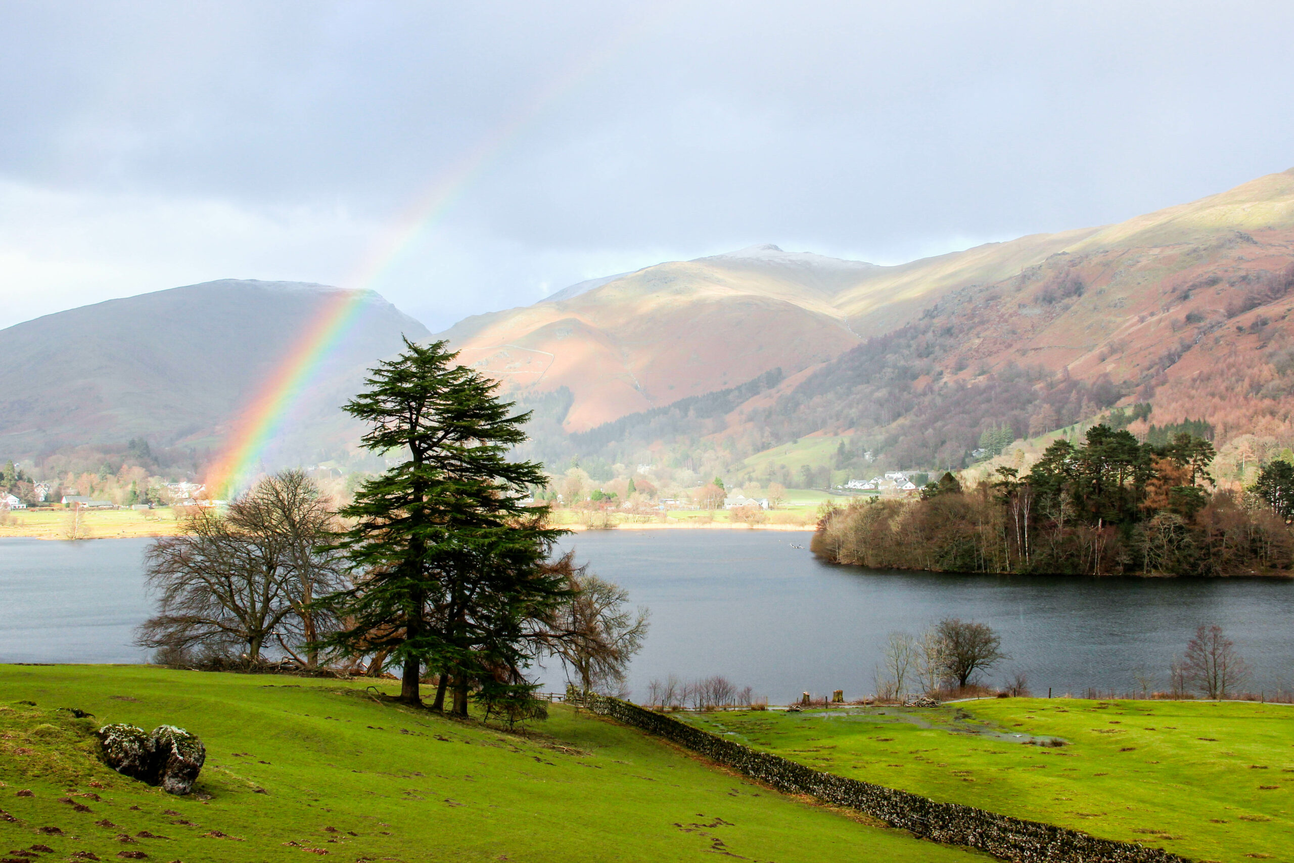 A rainbow rises above a tree, over a wide charcoal blue hued lake to the direction of orange and green landscape hills.
