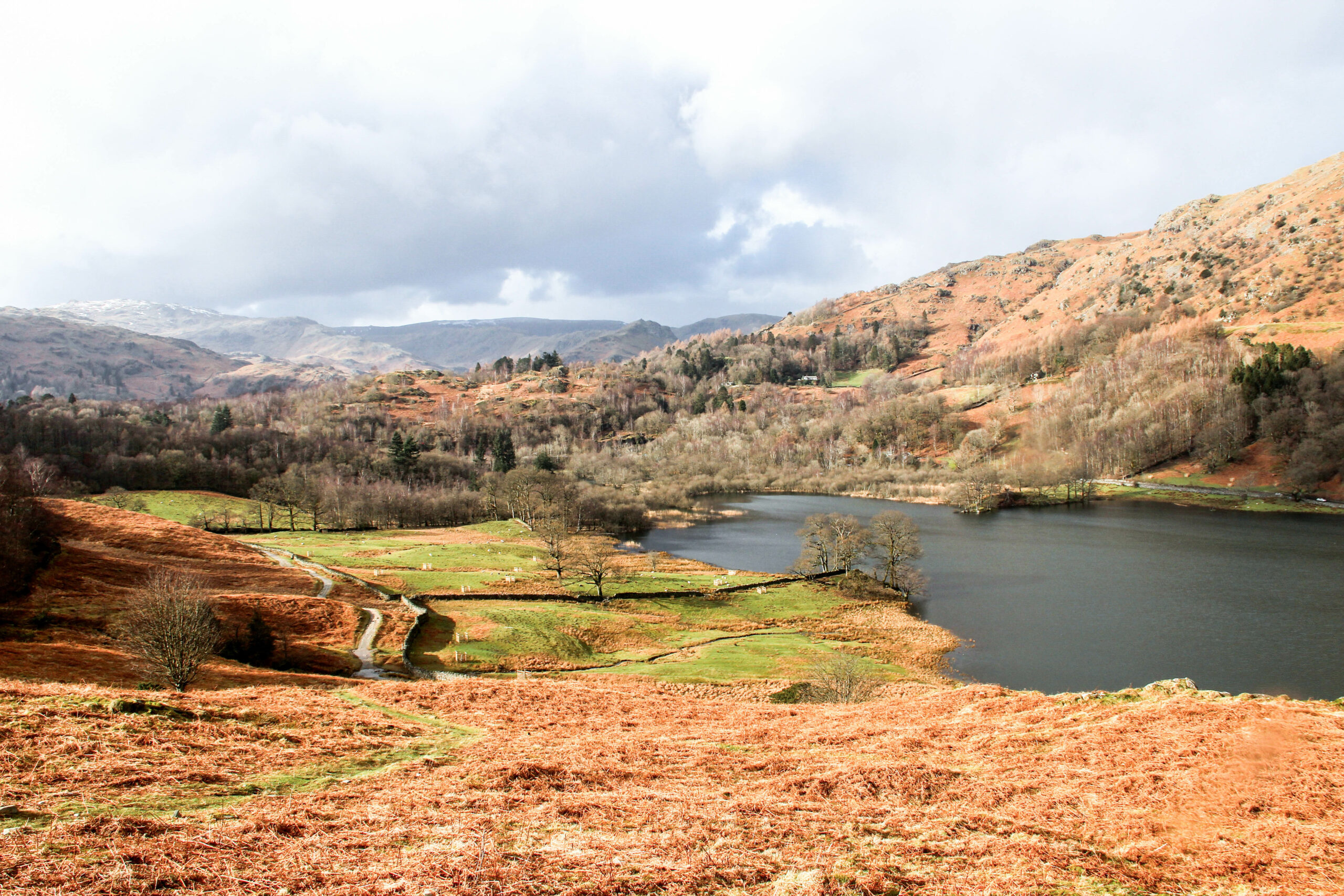 Far view of an orange field hiking path and green flat land net to a lake, backed by foliage covered undulating hills.