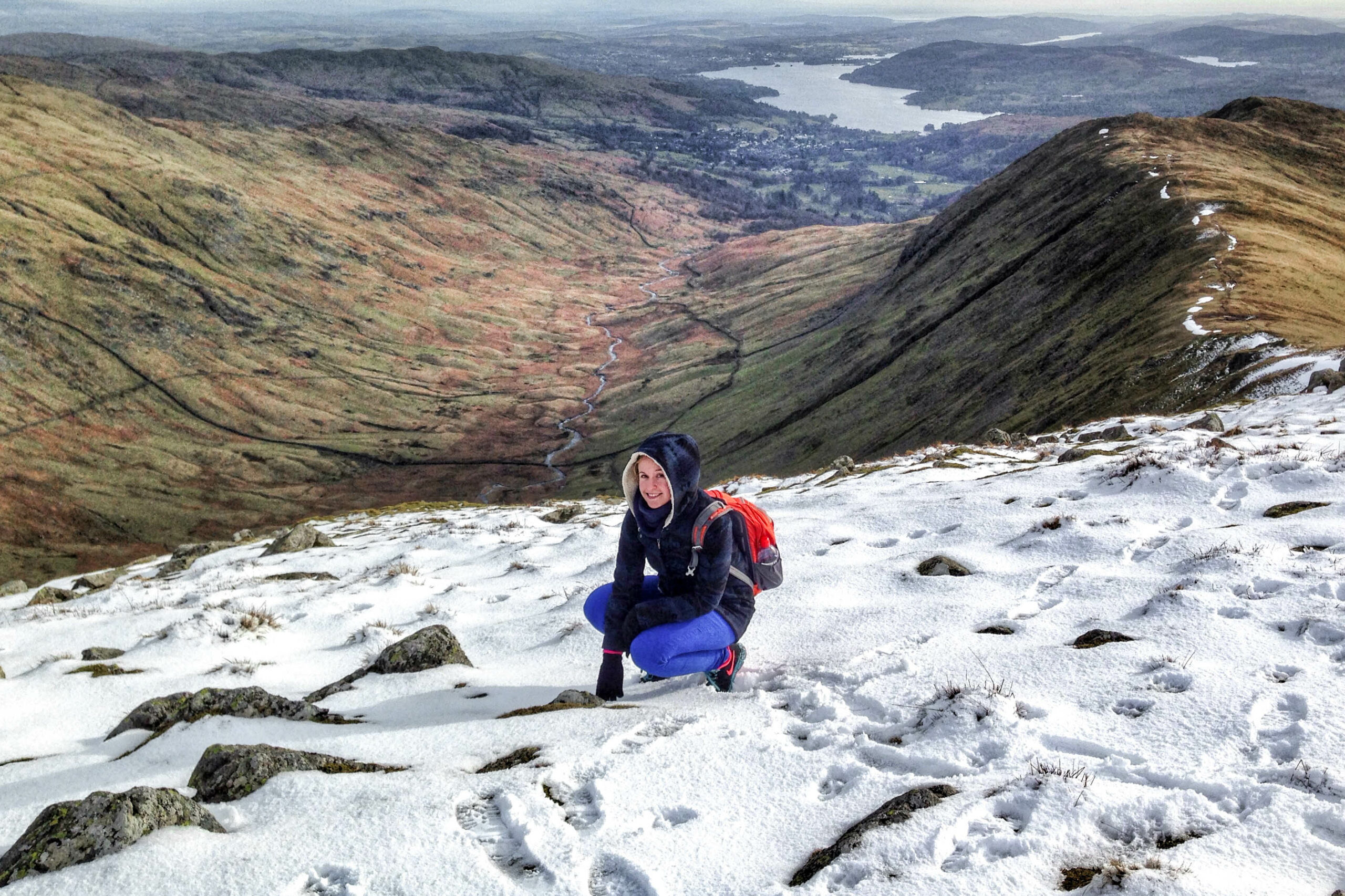 A woman dressed in blue and wearing a red backpack crouches down on a snow covered hill to a backdrop of a green valley and a distant lake.