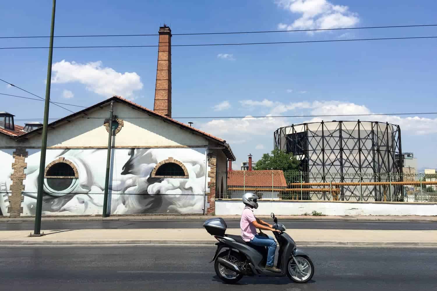 A person on a motorcycle rides past a modern triangular roofed building covered in street art, a golden bricked column and a round building with a metal and glass exterior - an industrial area of Athens. 