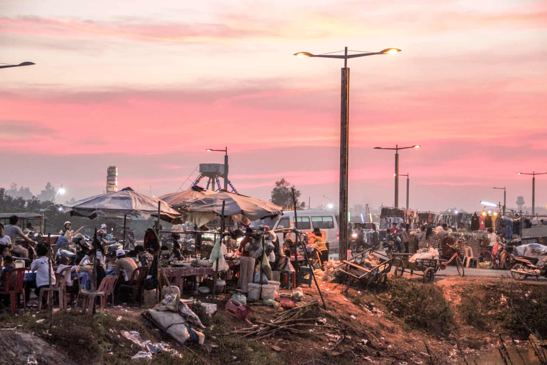 Behind a patch of land strewn with garbage and tall street lights, and a large group of people sit together on tables under umbrellas. A market stall with clothing is set up in the background. The sky is a mix of pink and yellow.