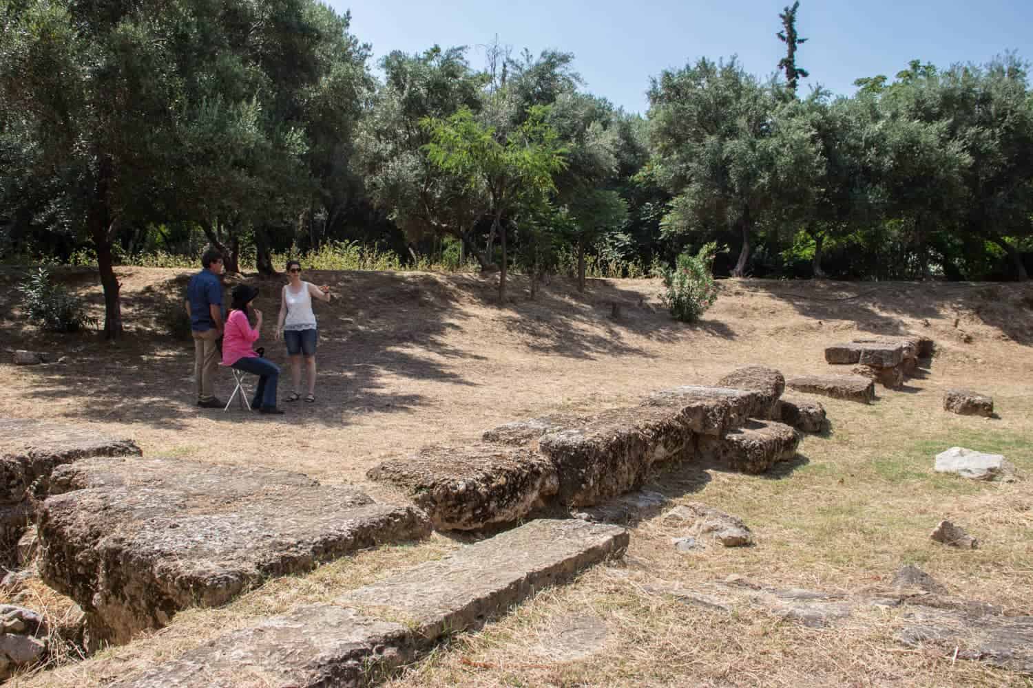 A woman in white points to a tree-back field of ruins as a man in blue and woman in pink look on - part of a Philosophy walking tours in Athens at Plato's Academy.