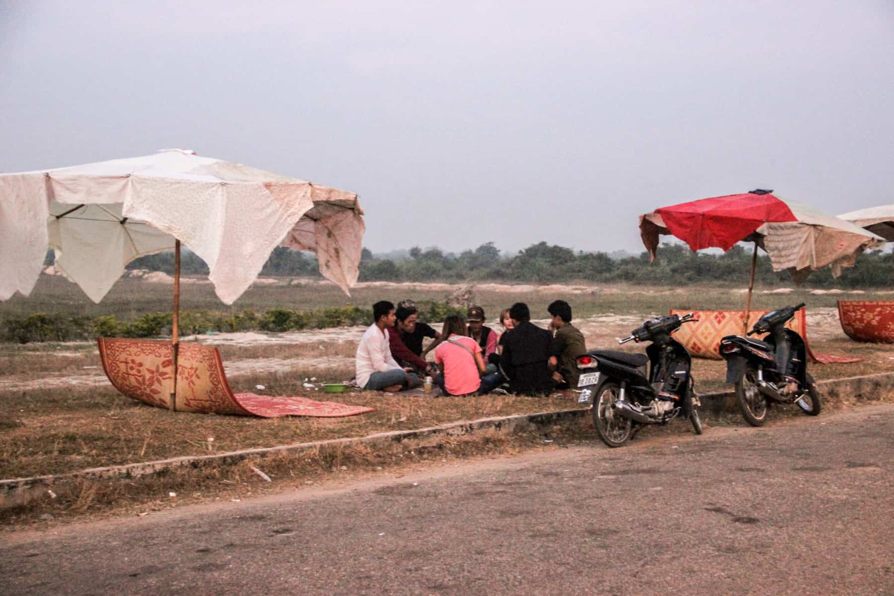 A group of eight people sit on road side grassland in between large umbrellas and in front of two motorbikes. In the background is flat green countryside. 