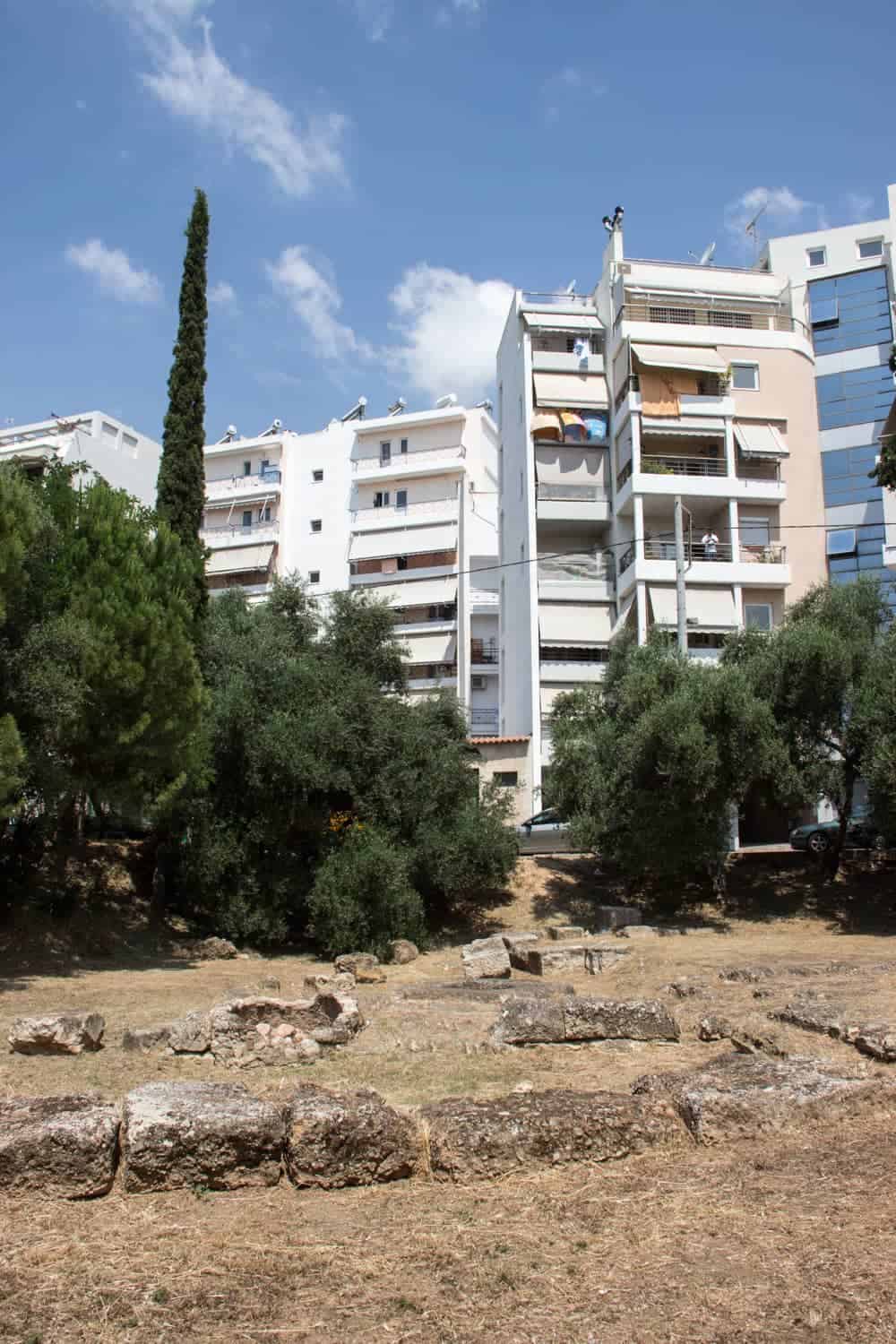 A white modern apartment building looms behind a tree-lined field full of stone ruins - part of Plato's Academy in Athens. walking tour