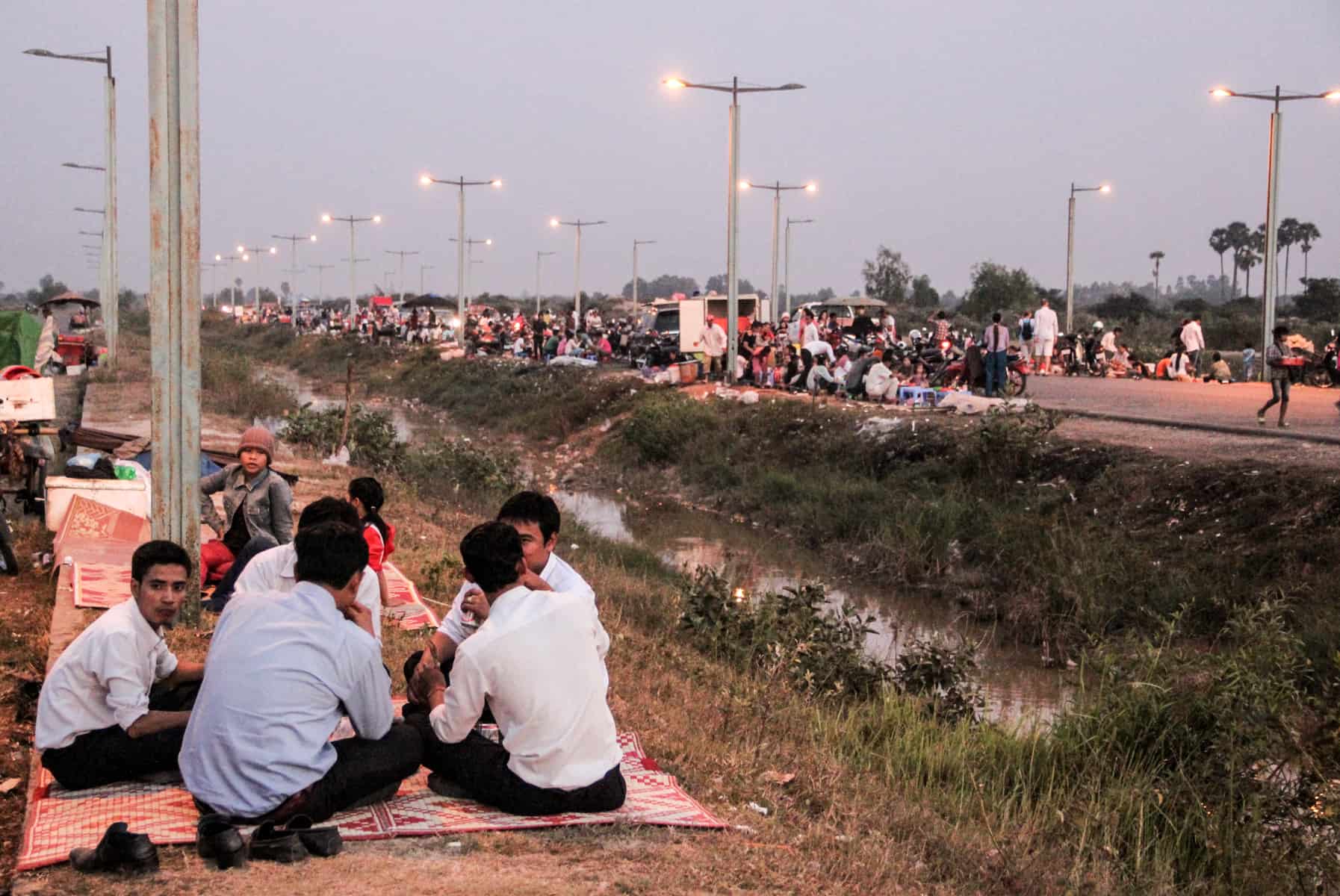 A group on men sit on a red blanket on grassland next to a narrow river. In the background dozens of people are sitting together on the other side of the road..