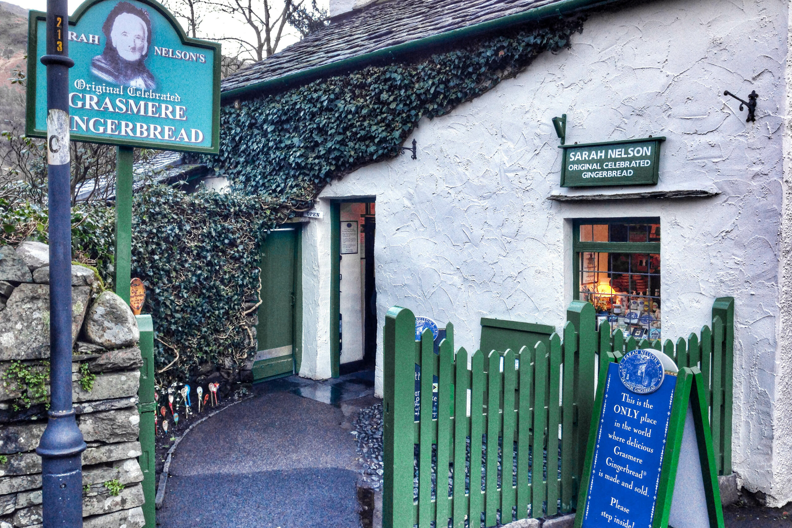 A white thatched cottage with a green gate and a sign reading 'Grasmere Gingerbread'. The sign above the window reads 'Sarah Nelson original celebrated gingerbread'.