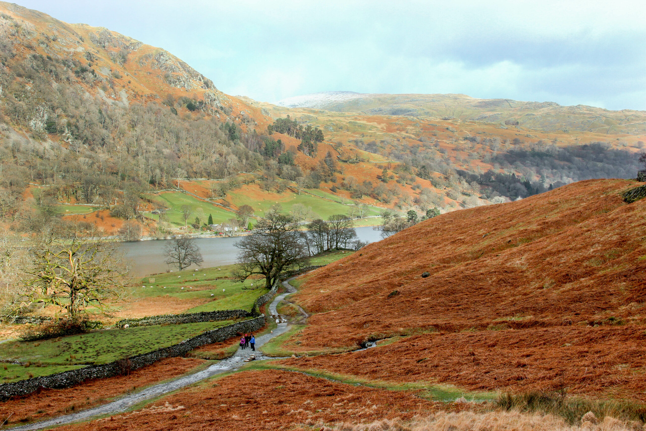 Slightly elevated view of a hiking path where a group of walkers have gathered. The path cuts through orange and green landscapes and leads towards the lakeside, backed by hills in the same hues.