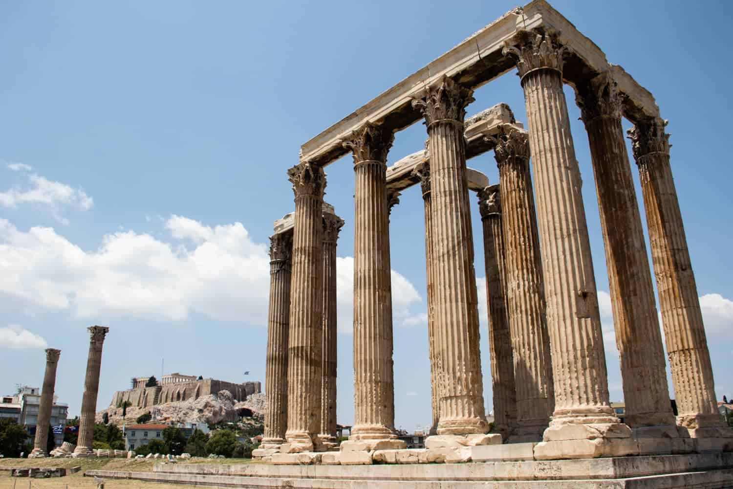 A tall, golden columned rectangular temple known as The Temple of Olympian Zeus - one of the ancient sites in Athens. The Acropolis can be seen in the background. 