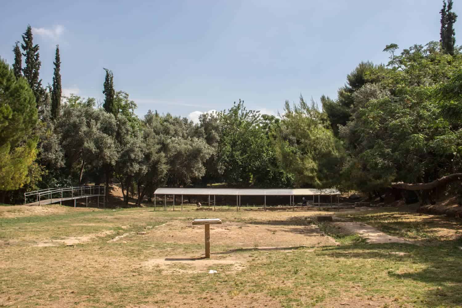 A bare field surrounded by trees, with a square indent of an old building in the middle and in the background, a line of ruins covered by a metal roof. Part Plato's Academy in Athen's. 