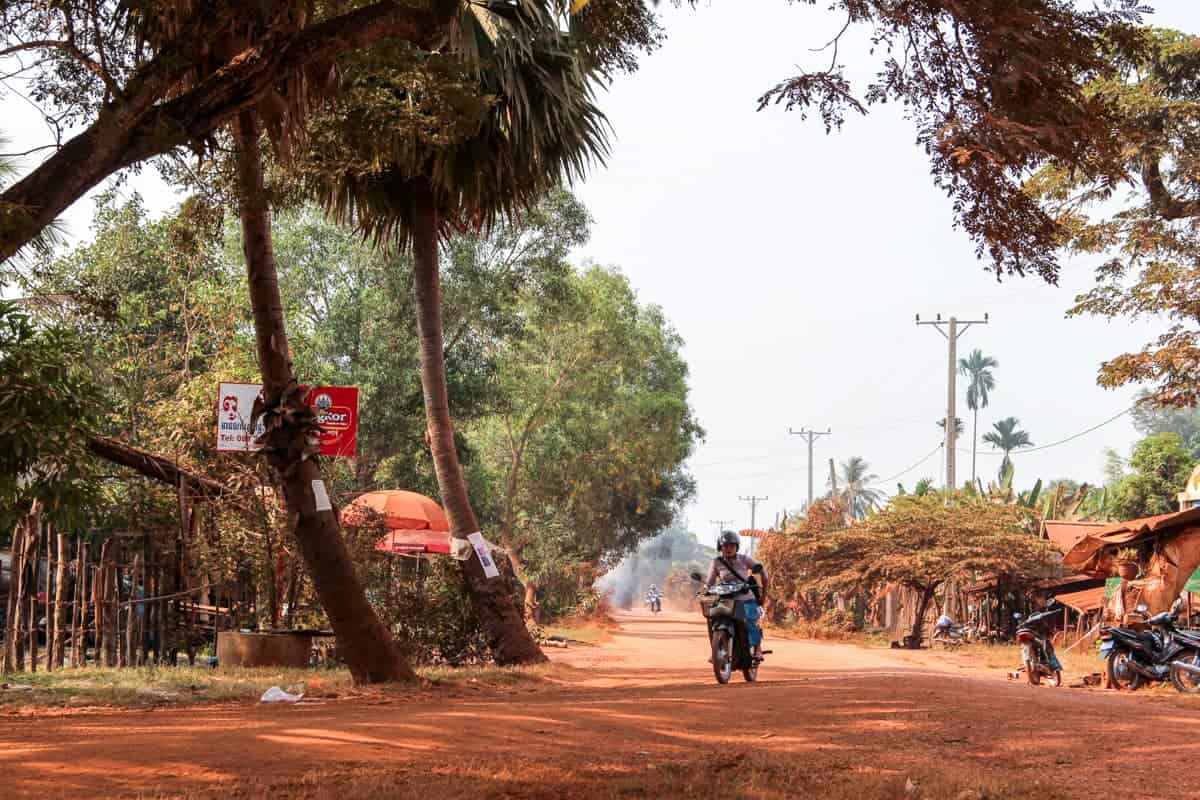 A man on a motorbike drives on an orange dusty road through a rural village in Cambodia