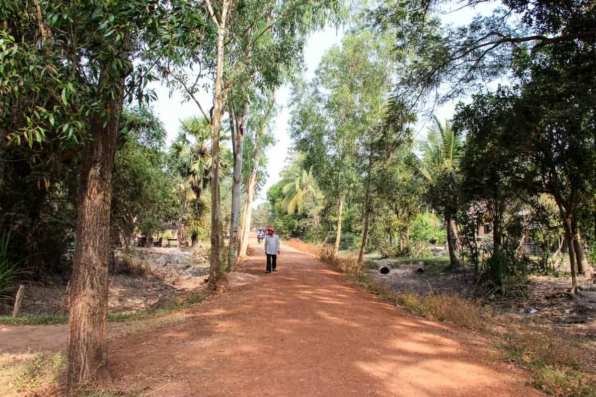 A man walks down an orange dirt road flanked on either side by lush green foliage and trees in a village in Cambodia