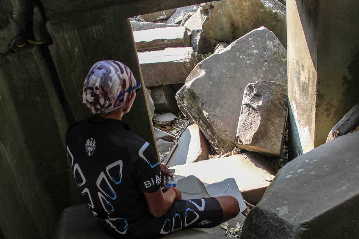 A man in biking clothing sits inside a hidden doorway filled with fallen stones at the jungle temple of Beng Mealea, Cambodia