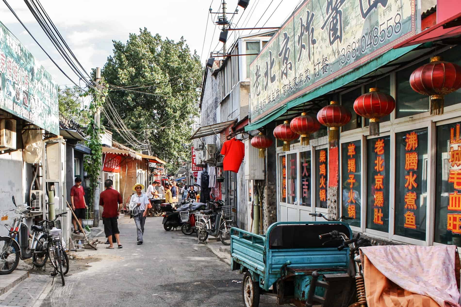 People surrounding street vendors in a Beijing Hutong near a shop with hanging red lamps. 