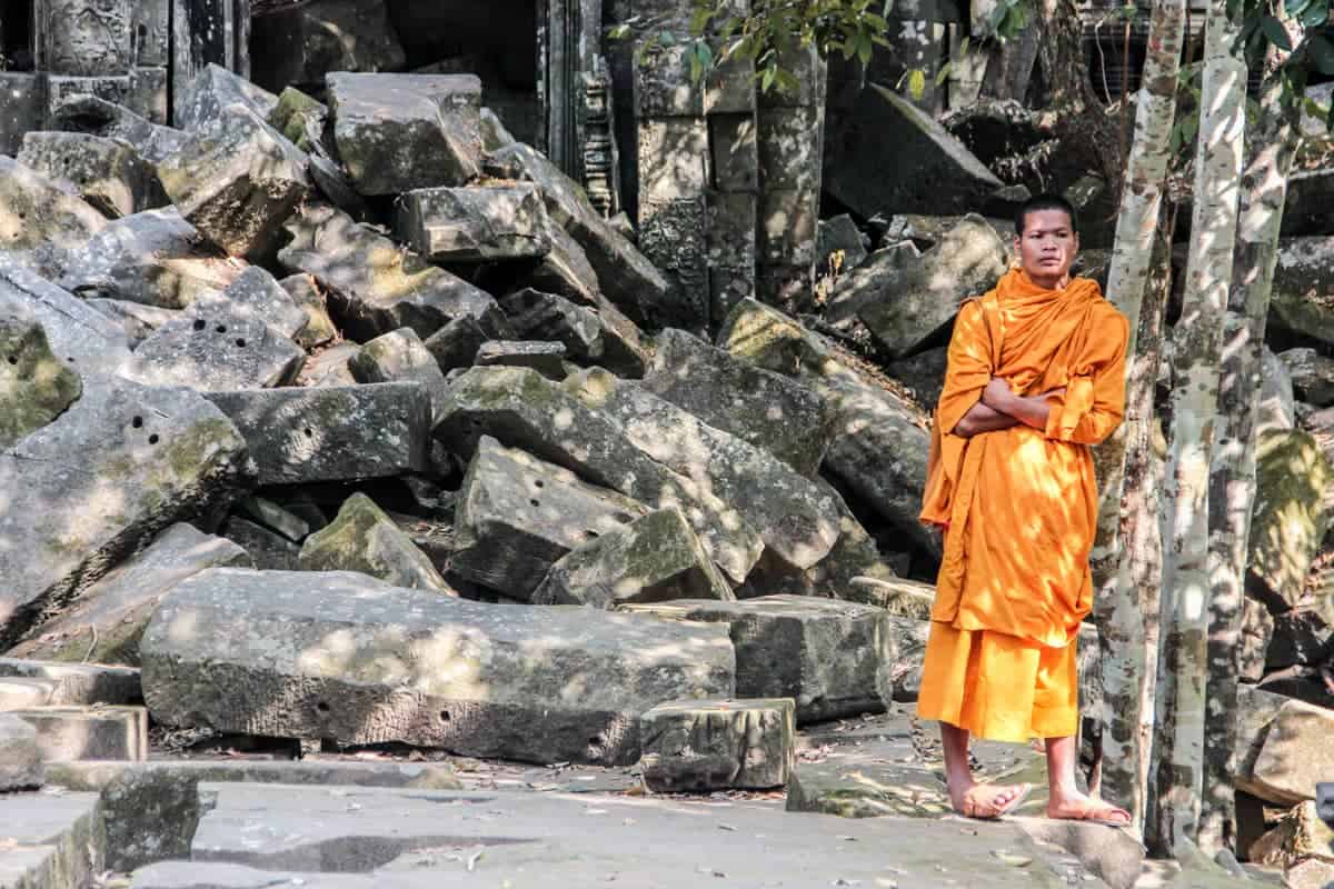 A monk in an ornage robe stands in front of a pile of fallen stones of Cambodia's jungle temple
